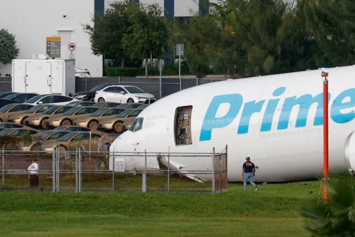 An Amazon Prime cargo jet sits off the runway behind a chain-link fence as an FAA official walks past.